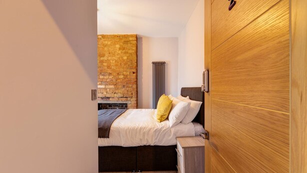 A bedroom with a brick wall, a bed with white bedding and a yellow pillow, a wooden door, and a radiator. - Bondgate Hotel East Midlands Airport (Derby)