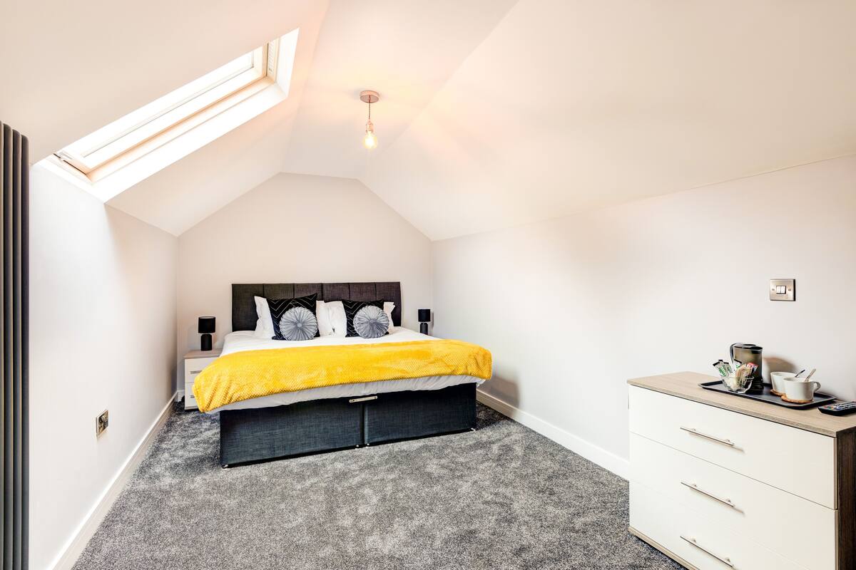 Une chambre avec un puits de lumière, un lit recouvert d’une couverture jaune, une table de chevet avec une lampe et une commode.