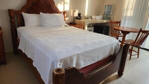 Un lit bien fait, avec une tête de lit en bois, une table de chevet avec une lampe, un petit bureau avec une chaise et un distributeur d’eau.