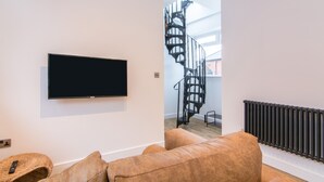 Una sala moderna con un televisor de pantalla plana montado en la pared, un sofá de cuero marrón, un reposapiés de mimbre y una escalera de caracol que conduce a un piso superior.