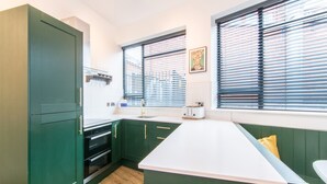 Cocina con armarios verdes, encimera blanca y ventana con persianas.