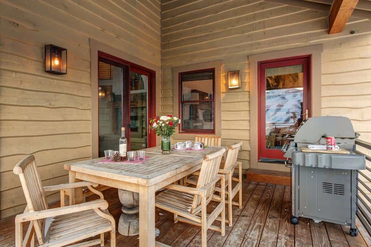 Una terraza de madera con una mesa, sillas, una parrilla y un ventanal con vistas.