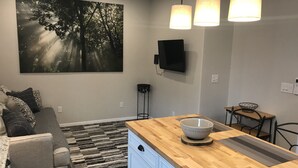 A modern kitchen with a wooden countertop, a dining area with a small table and chairs, a gray sofa with patterned cushions, a large abstract painting on the wall, and a ceiling fan.