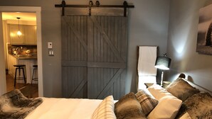 A bedroom with a bed, a large wooden barn door, a desk with a lamp, and a kitchen visible through an open door. - Cozy Condo in the Heart of Mont Tremblant Resort                (Mont-Tremblant)