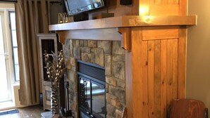 A cozy living room with a stone fireplace, a mounted television, and a wooden chair. - Cozy Condo in the Heart of Mont Tremblant Resort                (Mont-Tremblant)