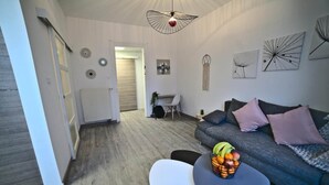 A modern living room with a grey sofa, a small round table with fruit, and a white chair. There is a dining area in the background with a table and chairs. - -- Love Life -- (Mulhouse)