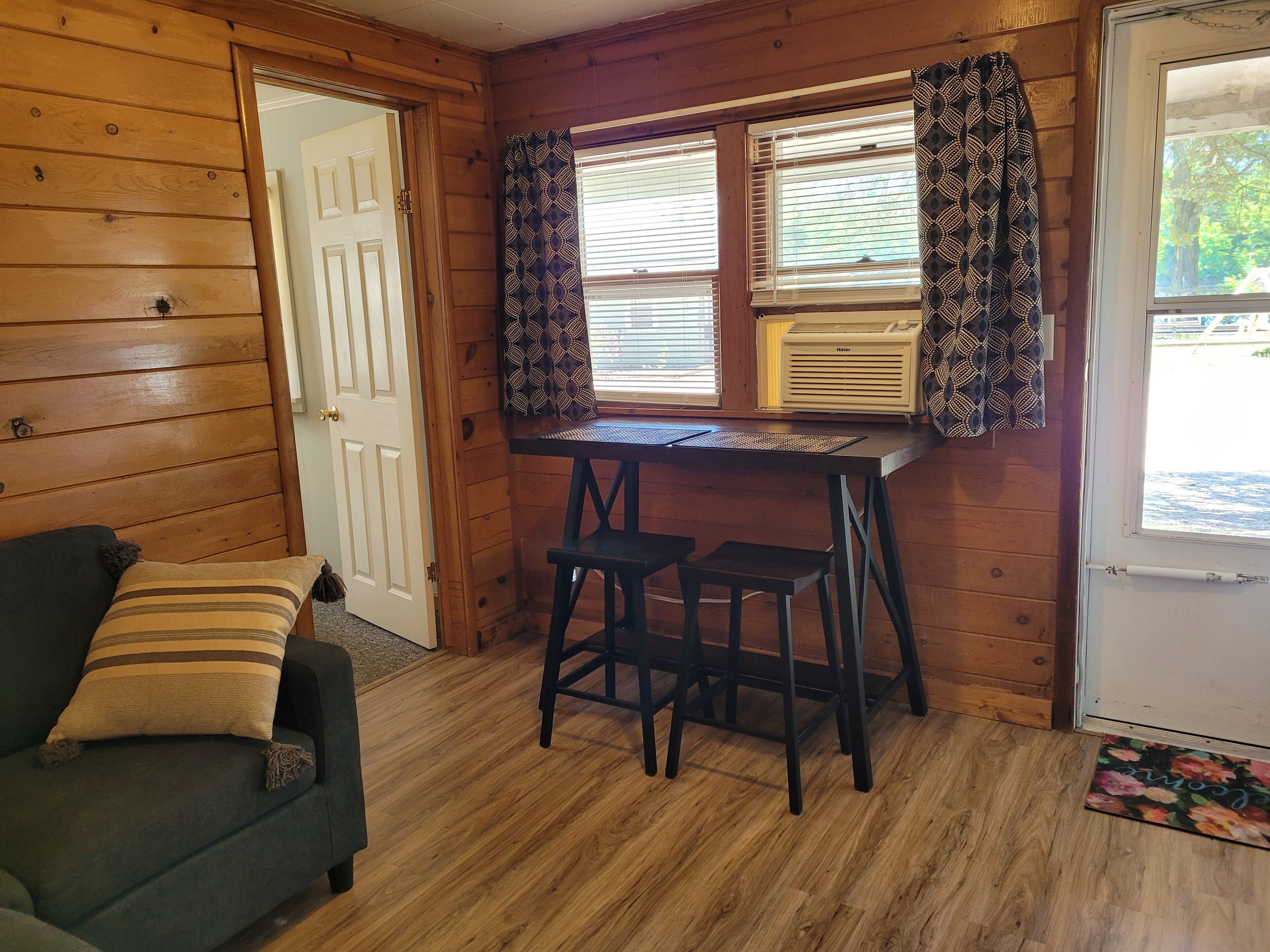 Una habitación acogedora con paredes de madera, una pequeña mesa con dos taburetes, una puerta y una ventana con cortinas estampadas.