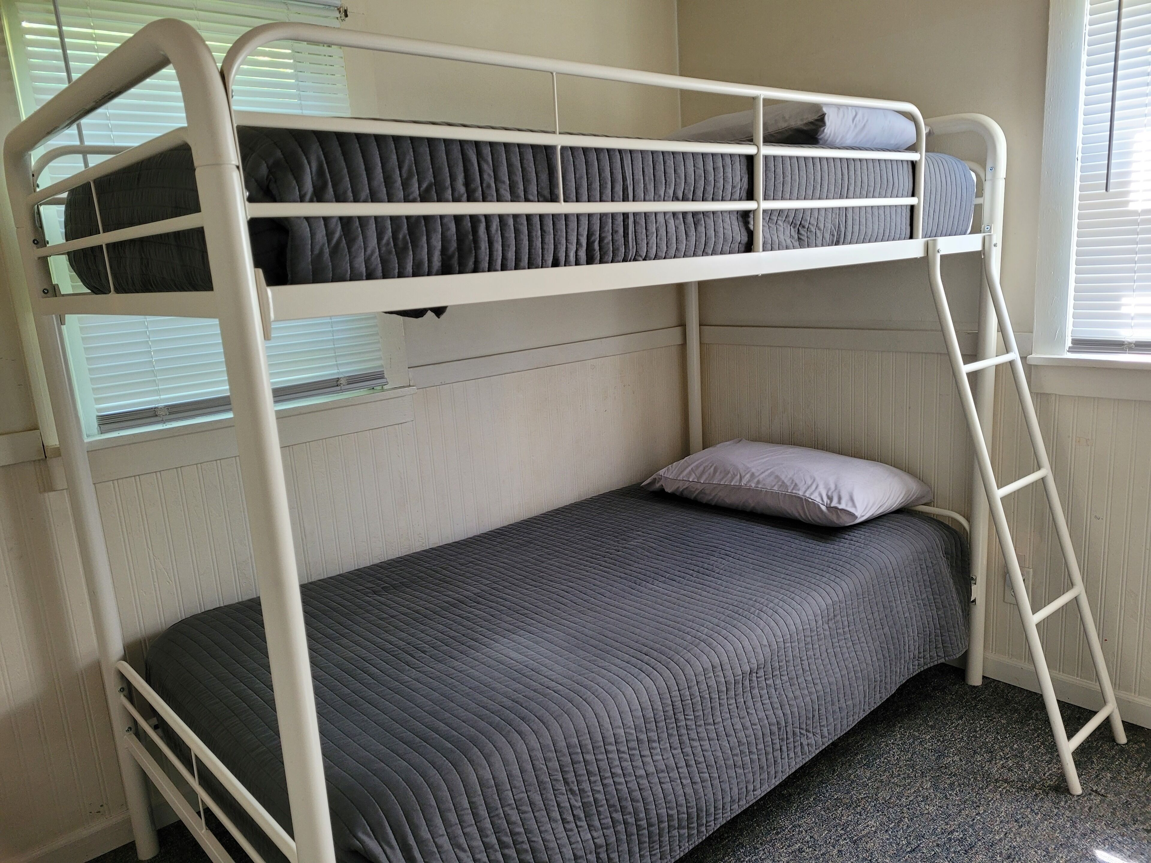 A bunk bed with a ladder, a single bed below, and a window with blinds.