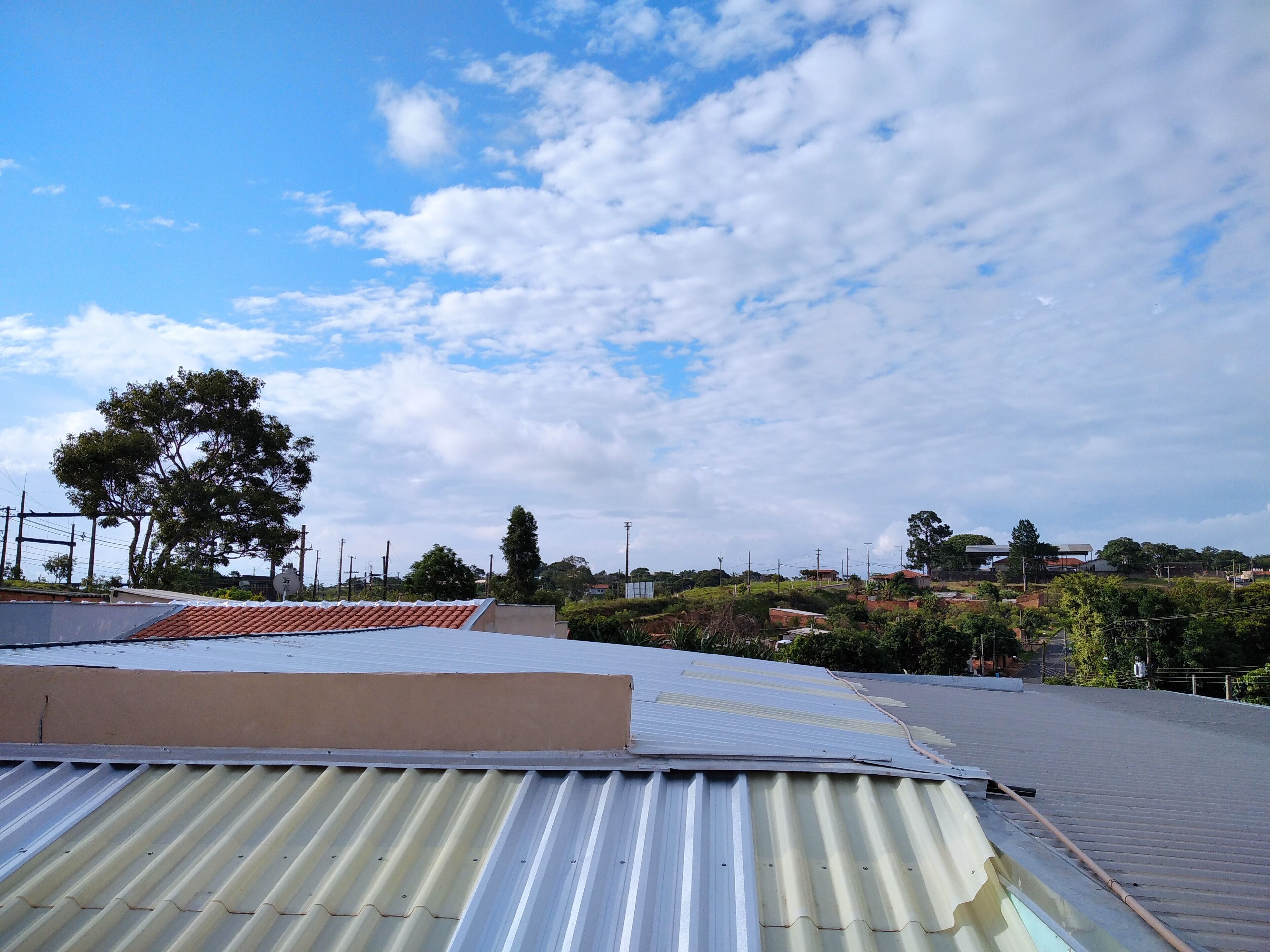 A rooftop with corrugated metal roofing, a large tree, and a distant view of a residential area with houses and greenery.