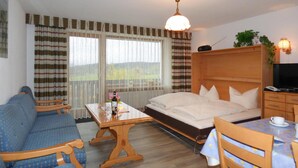 Una habitación con cama, sofá, mesa con una botella y vasos, televisor y ventana con cortinas.