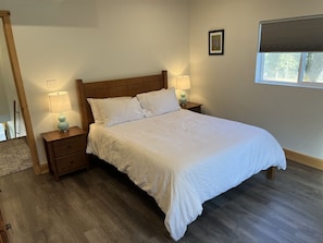 A bedroom with a wooden bed, two bedside tables with lamps, a window with blinds, and a framed picture on the wall.