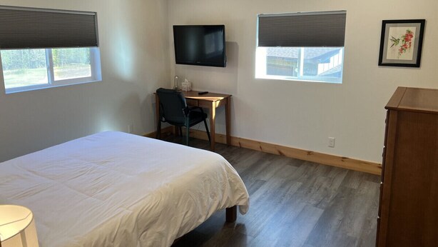Un dormitorio con una cama, un escritorio, una silla, un televisor y dos ventanas con persianas.