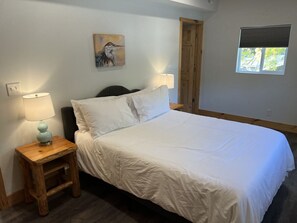 A bedroom with a bed, two bedside lamps, a wooden nightstand, a painting on the wall, and a window with blinds.