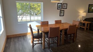 Een eetgedeelte met een houten tafel en stoelen, een hanglamp en een raam met uitzicht op bomen.