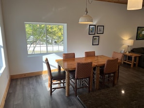 A dining area with a wooden table and chairs, a hanging light fixture, and a window with a view of trees.