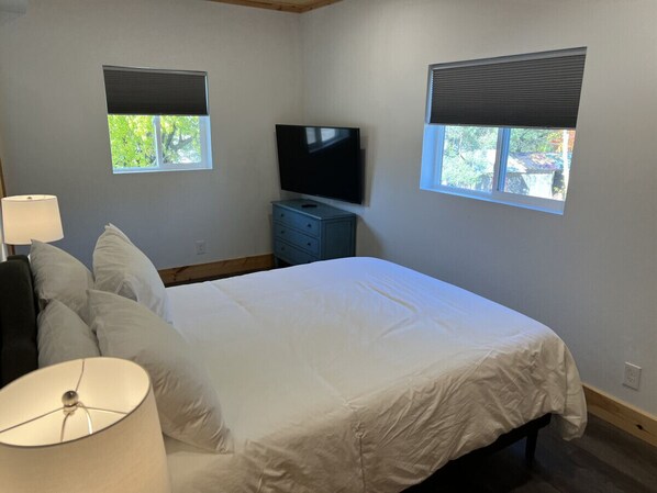 Ein Schlafzimmer mit einem Bett, einem Fernseher, zwei Fenstern mit Jalousien, einer Lampe und einer Kommode.