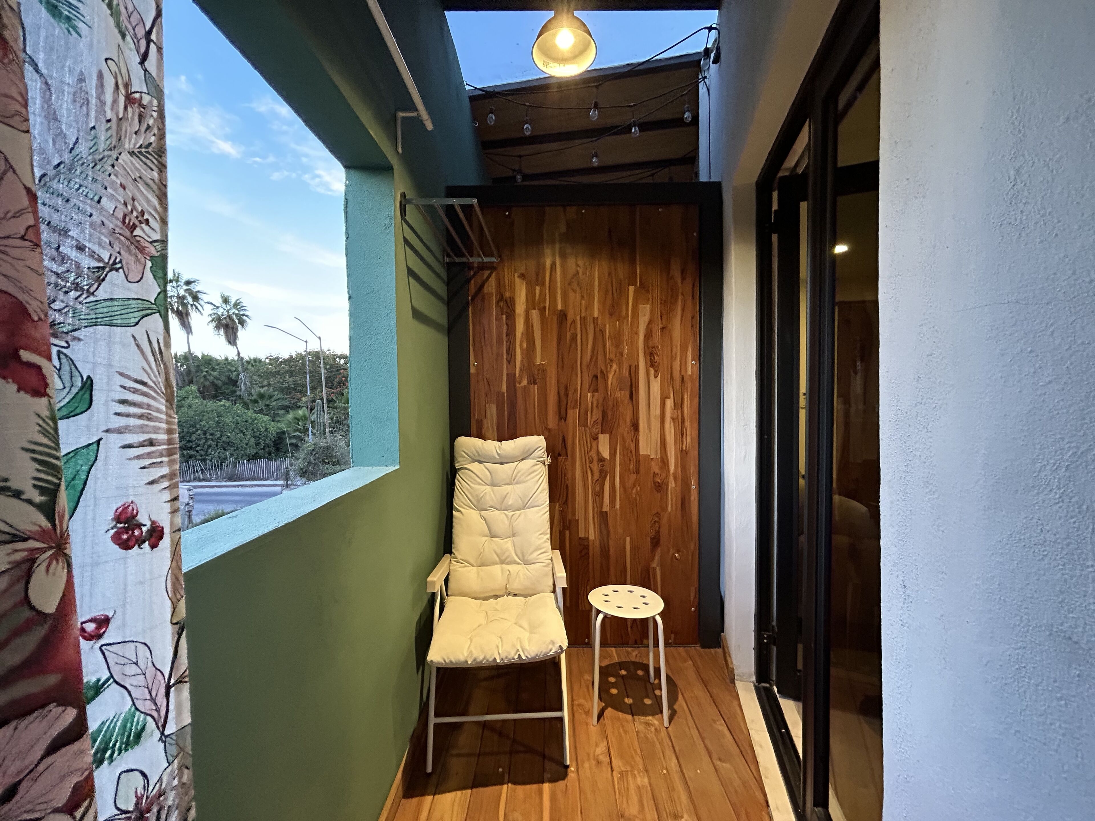 Un balcón con una silla acolchada y un taburete pequeño, suelo de madera, una pared verde y una puerta de madera.