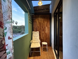 Un balcón con una silla acolchada y un taburete pequeño, suelo de madera, una pared verde y una puerta de madera.