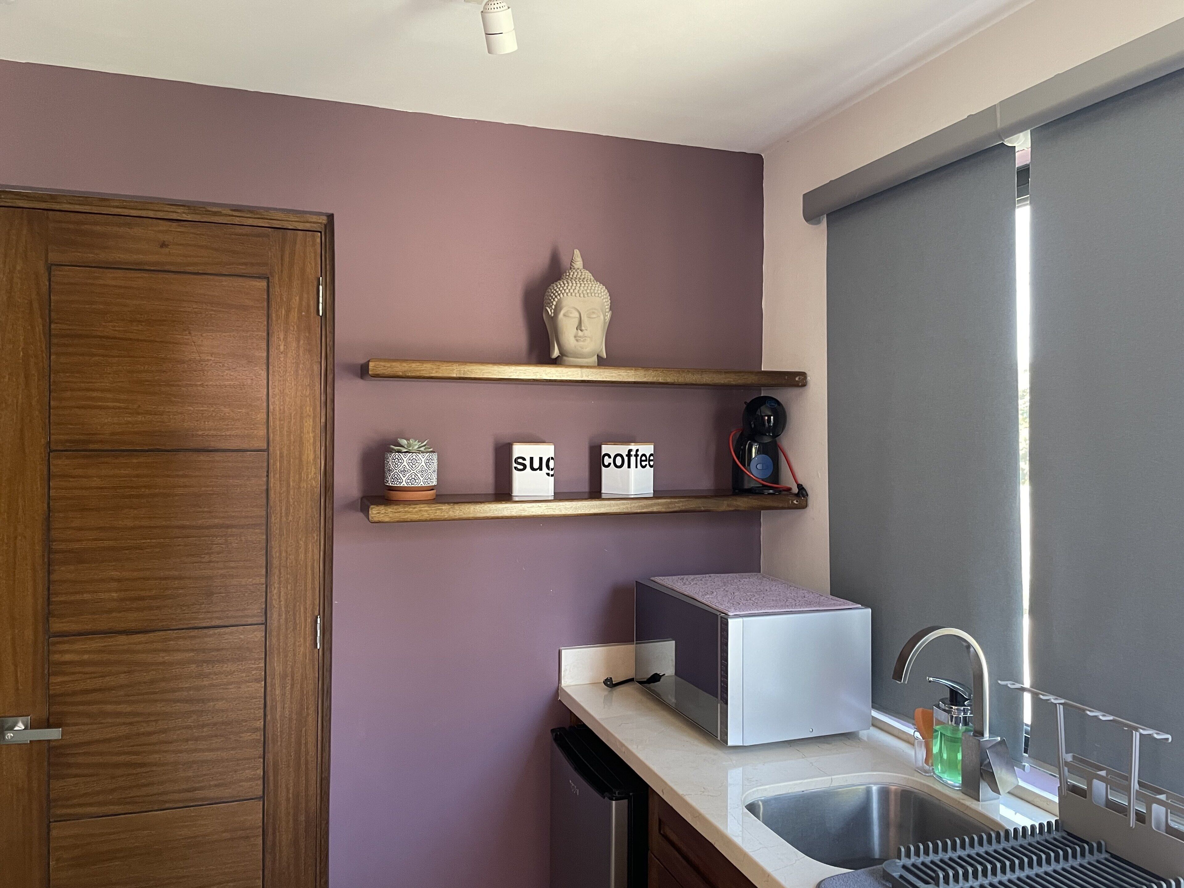 Una cocina con armarios de madera, microondas integrado, cafetera y ventana con persiana.