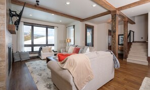A spacious living room with a large window, wooden beams, a fireplace, and a staircase.