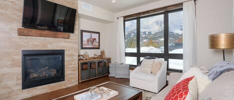 Un salon avec une cheminée, un téléviseur fixé au mur, une grande fenêtre donnant sur les montagnes, un canapé blanc et une table basse en bois.