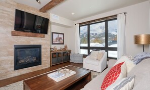 A living room with a fireplace, a mounted TV, a large window with a mountain view, a white sofa, and a wooden coffee table.