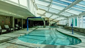 Une piscine intérieure dotée d’un toit en verre, d’un sol en pierre et de chaises longues disposées le long des parois.