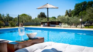 A swimming pool with a clear glass and bowl on a wooden table, surrounded by greenery and a thatched umbrella.