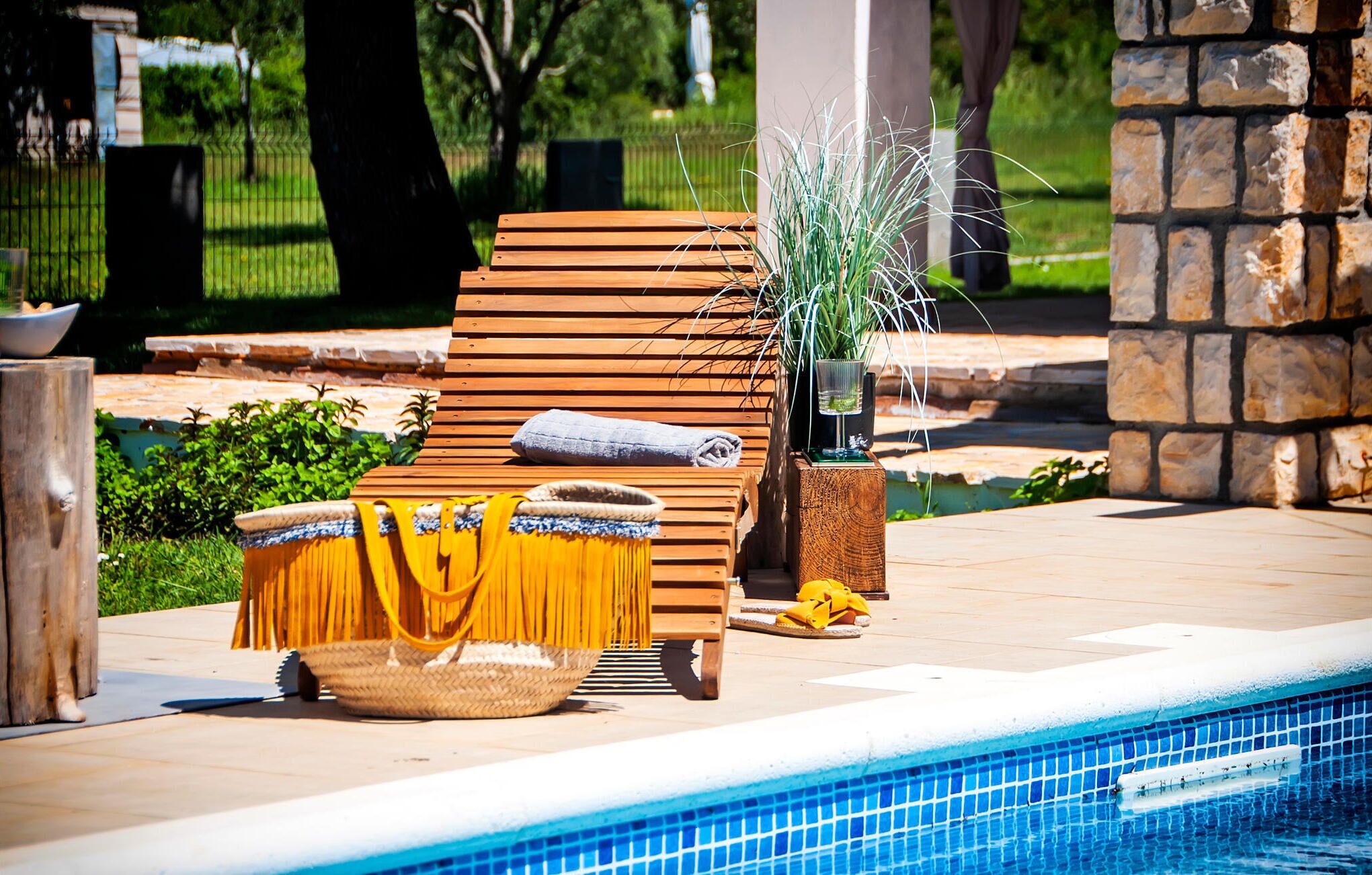 A wooden lounge chair with a towel and a yellow towel on a poolside deck.