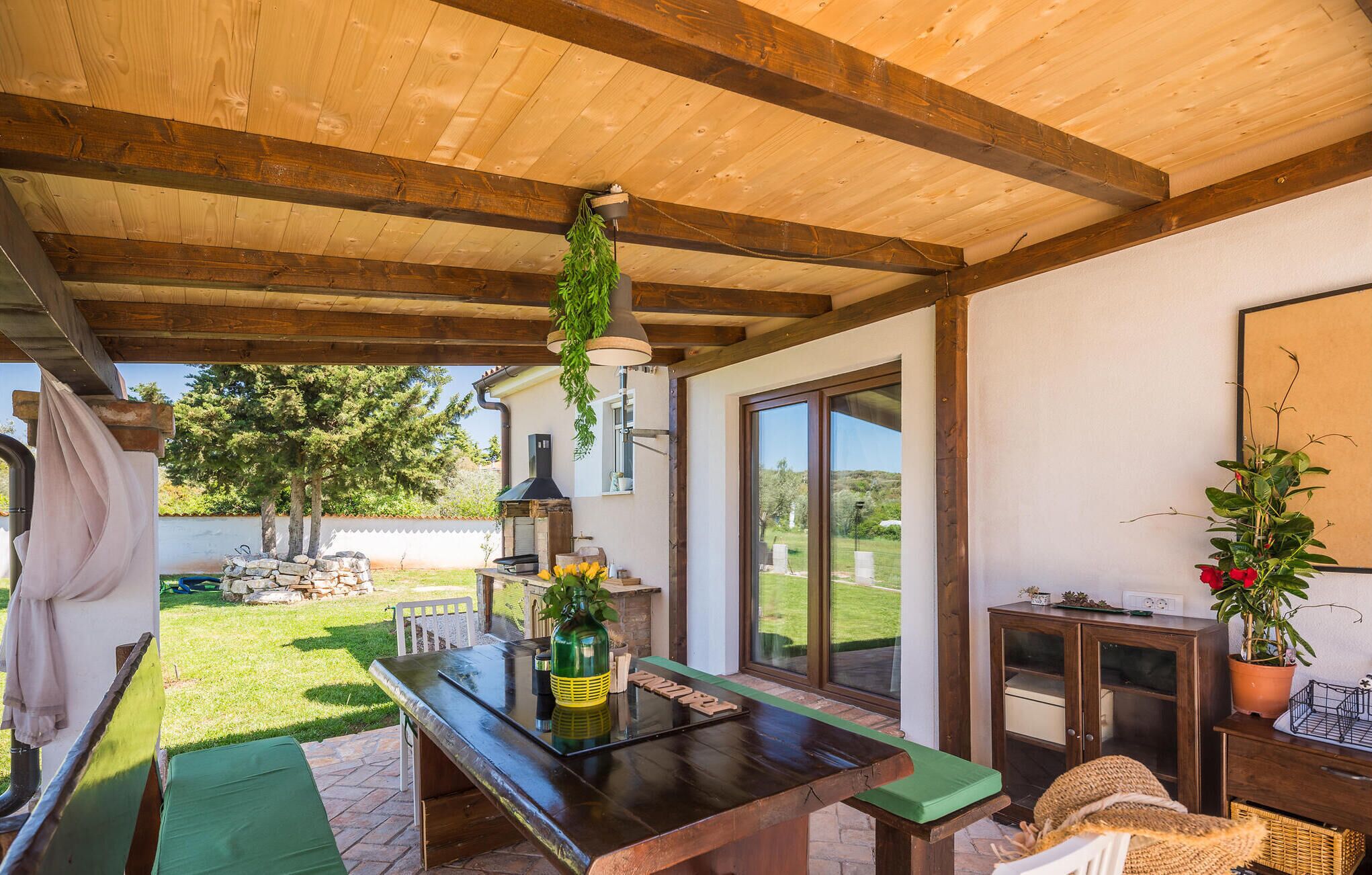Un espace extérieur abrité, doté d’un plafond en bois, d’une table avec une bouteille et des verres, d’une cheminée en pierre et d’une vue sur le jardin.