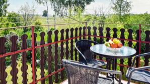Un balcón con una silla y mesa de mimbre, un frutero con fruta, y vistas a zonas verdes y árboles.