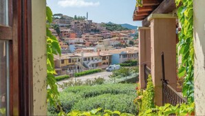A view from a balcony with a wooden railing and a window frame, overlooking a hillside with colorful buildings and a clear sky. - Rokkaria by Interhome (Villasimius)
