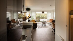 Uma sala de estar moderna com sofá, cadeiras, mesa de centro e área de jantar. Grandes janelas oferecem luz natural e vista para o verde lá fora.