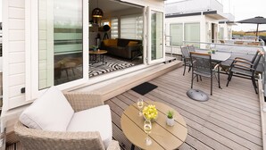 Um terraço com área de estar externa, uma mesa com uma cesta de frutas e taças de vinho, e vista do interior através de grandes portas de vidro.