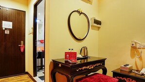 A hotel room with a wooden door, a mirror, a bedside table with a red towel, a bedside lamp, and a telephone.