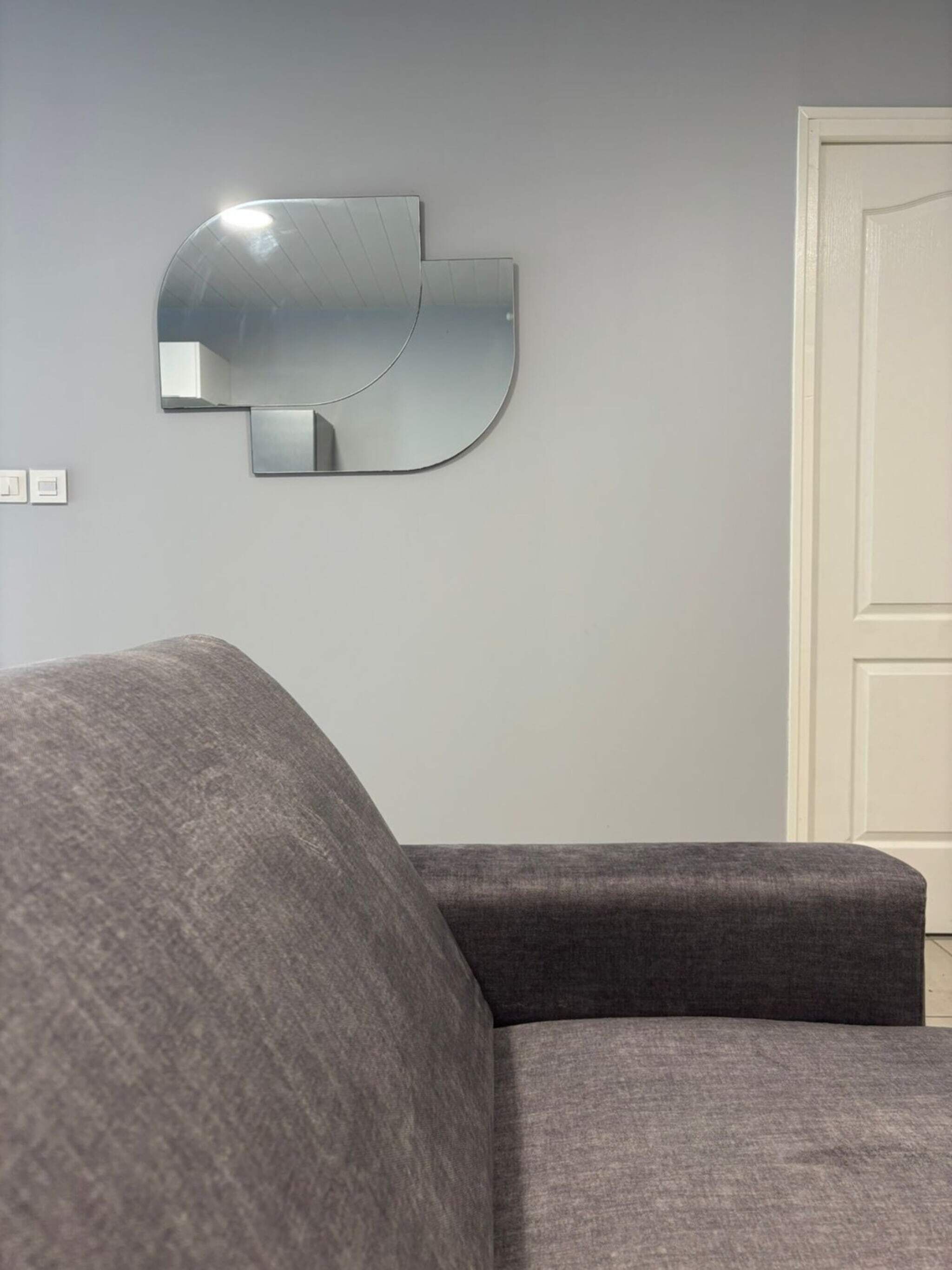 Un salón moderno con un sofá gris, un espejo montado en la pared y una puerta cerrada.