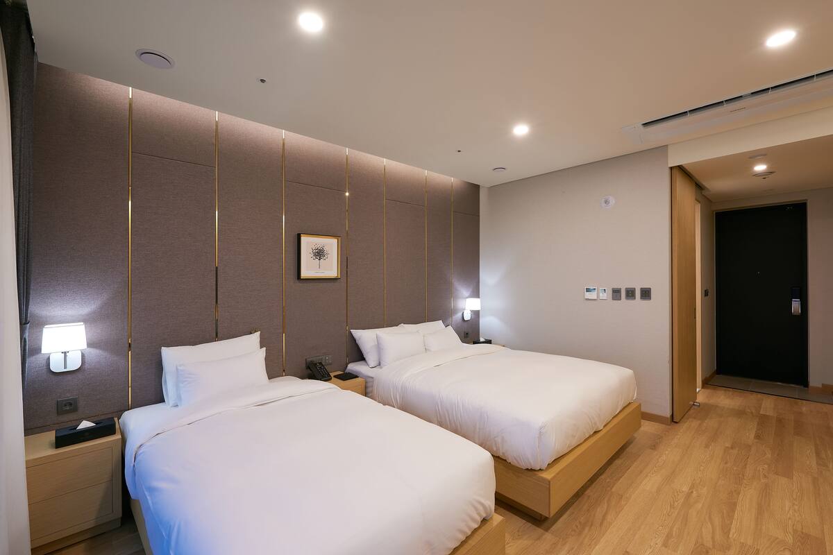 Una habitación de hotel con dos camas, una mesita de noche con lámpara, un cuadro enmarcado en la pared y una puerta que da a otra habitación.