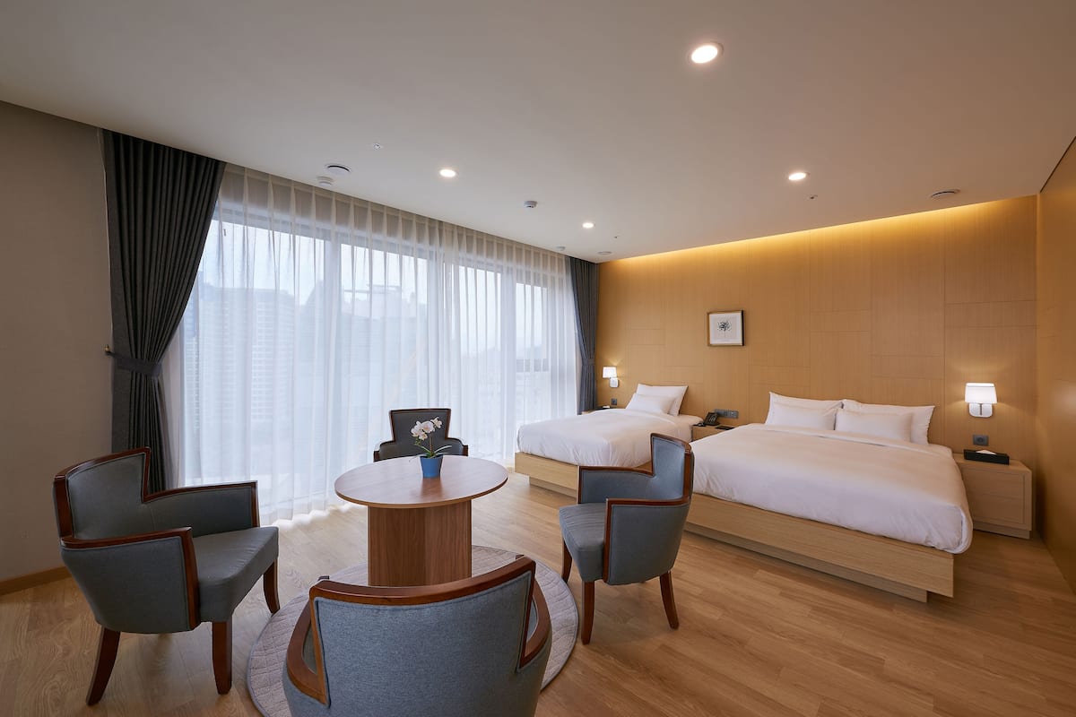 Una habitación de hotel con una cama, dos mesitas de noche, una mesa redonda con sillas y un amplio ventanal con cortinas.
