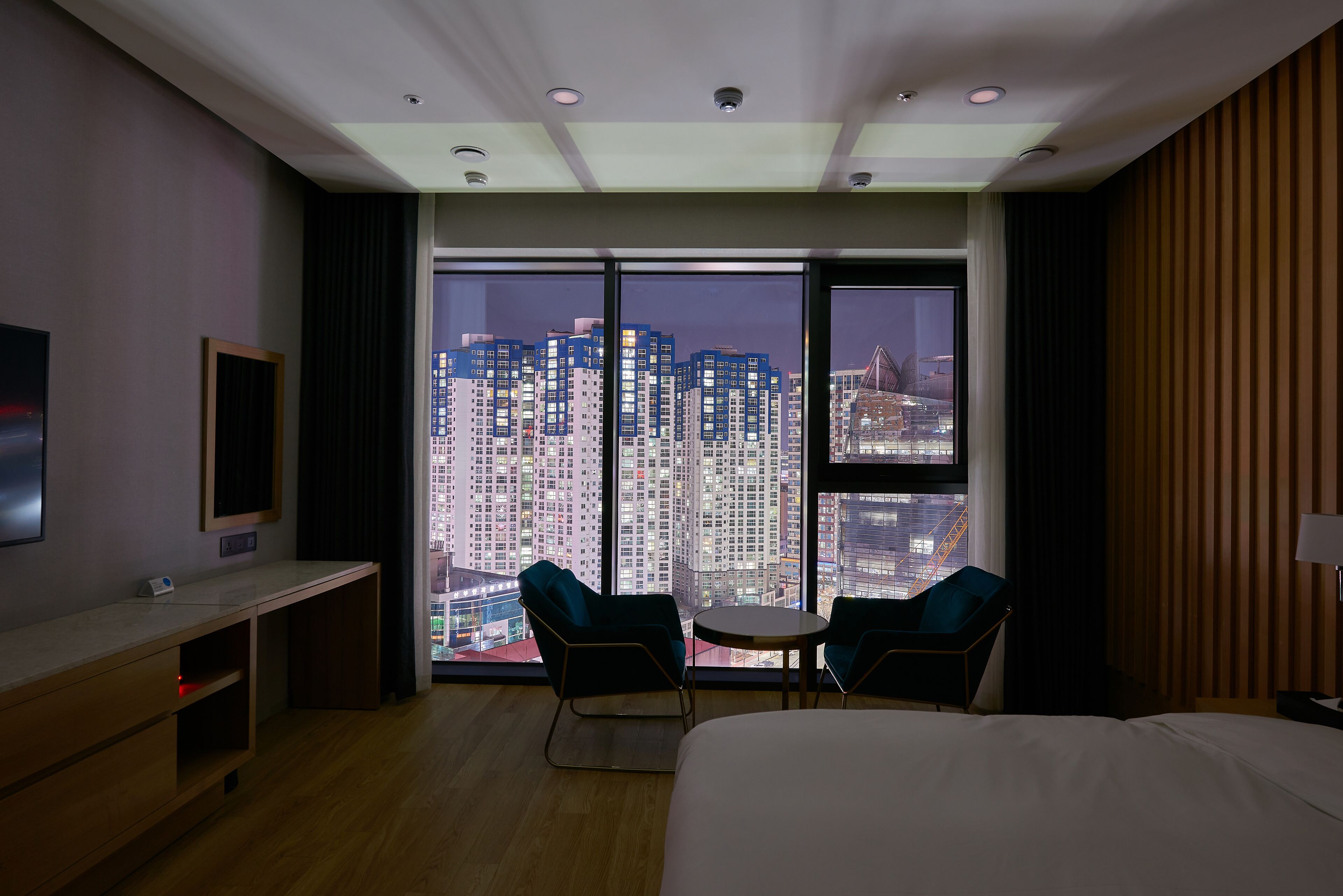 A hotel room with a large bed, a desk, two chairs, a TV, and a city view through floor-to-ceiling windows.