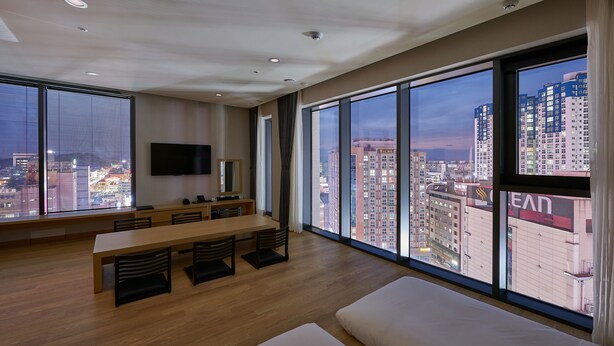 Een moderne hotelkamer met een groot raam met uitzicht op de stad, een zithoek met een lange tafel en stoelen, en een flatscreen televisie aan de muur.