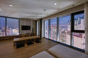 A modern hotel room with a large window offering a city view, a dining area with a long table and chairs, and a flat-screen TV mounted on the wall. - CS Hotel (Gwangju)