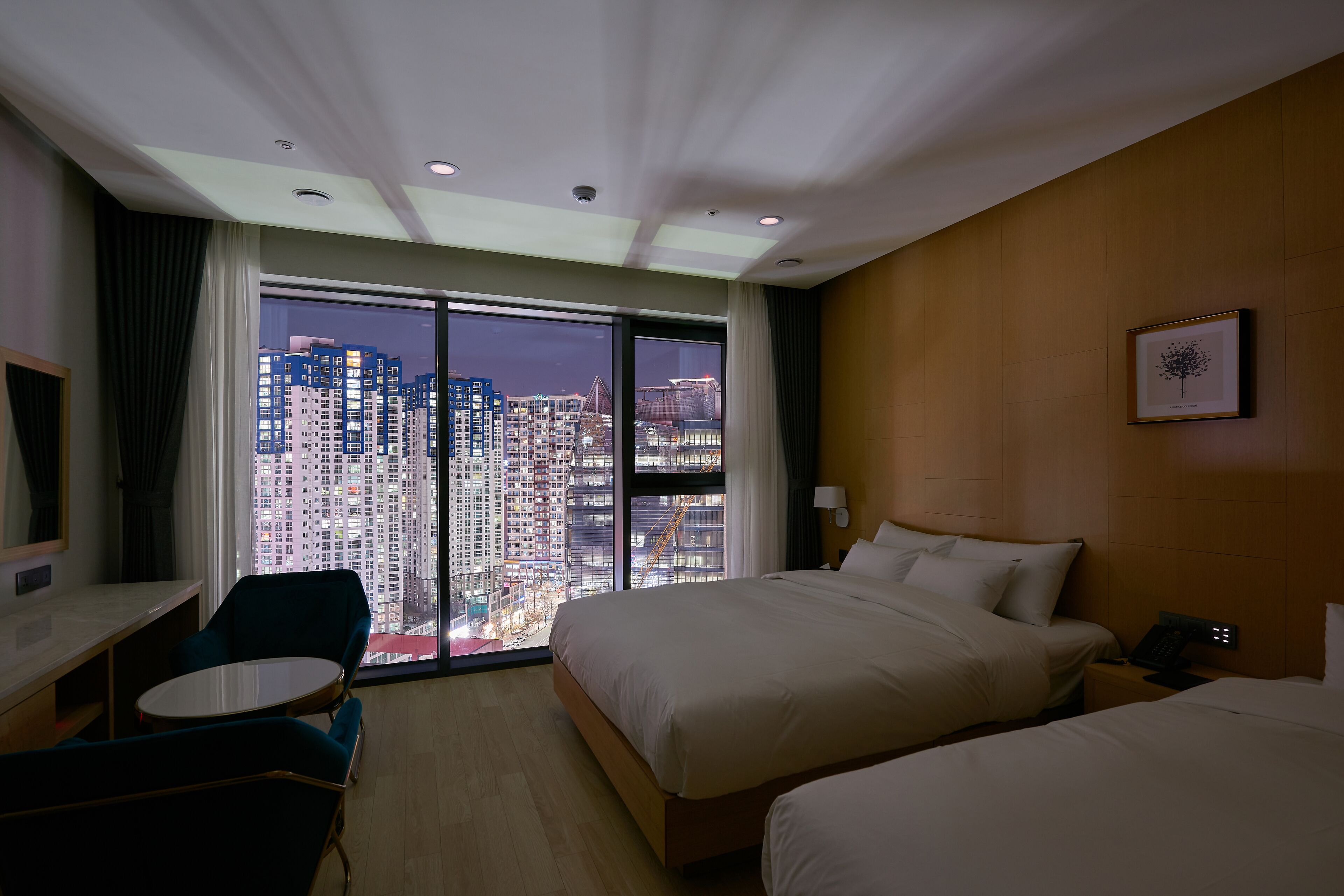 Habitación de hotel con una cama grande, una cama más pequeña, un escritorio y una silla. La habitación tiene vista a edificios altos.