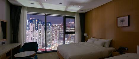 Habitación de hotel con una cama grande, una cama más pequeña, un escritorio y una silla. La habitación tiene vista a edificios altos.