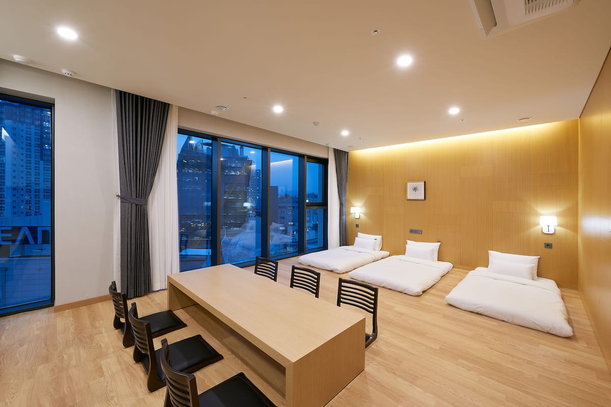 Habitación de hotel moderna con un gran ventanal, suelo de madera, una mesa de comedor central y dos camas con ropa blanca.