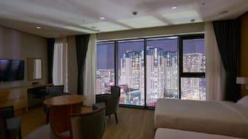 Ein Hotelzimmer mit großem Fenster und Blick auf die Stadt, einem Bett, einem Schreibtisch mit Stuhl und einem Fernseher.