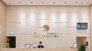 Een receptiebalie van een hotel met een modern design, een digitaal scherm erboven en een potplant aan de rechterkant.