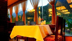A dining area with a table set for two, a view of mountains through large windows, and a chandelier. - Pine Spring Hotel (Anantnag)