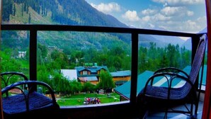 A room with a large window offering a view of a mountainous landscape and a small building with a green roof. - Pine Spring Hotel (Anantnag)
