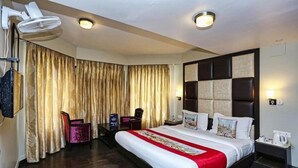 A hotel room with a large bed, a desk, a chair, a fan, and curtains. - Pine Spring Hotel (Anantnag)