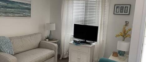 A living room with a striped sofa, a teal armchair, a TV on a white cabinet, a lamp, and a painting on the wall.